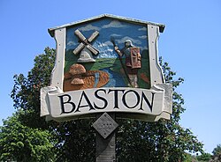 Closeup of village sighn, with a blue sky and green tree in the background. The sign is framed like a medieval barn, around a low relief carving, painted in realistic colours, featuring a stream with a Roman soldier on the right and an 18th century smock mill on the left.