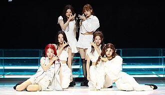 A group of six women performing onstage, while appearing in white streetwear clothing